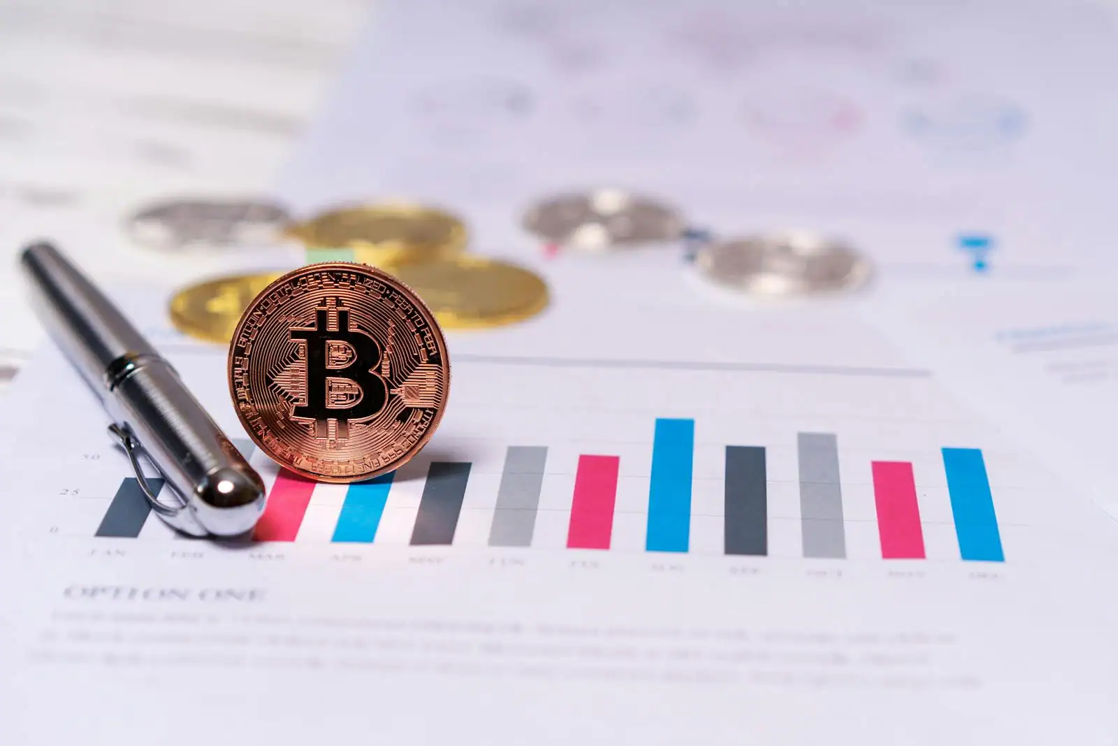 Pièce physique à l'effigie du bitcoin posée sur un graphique en barres, avec d'autres pièces et un stylo