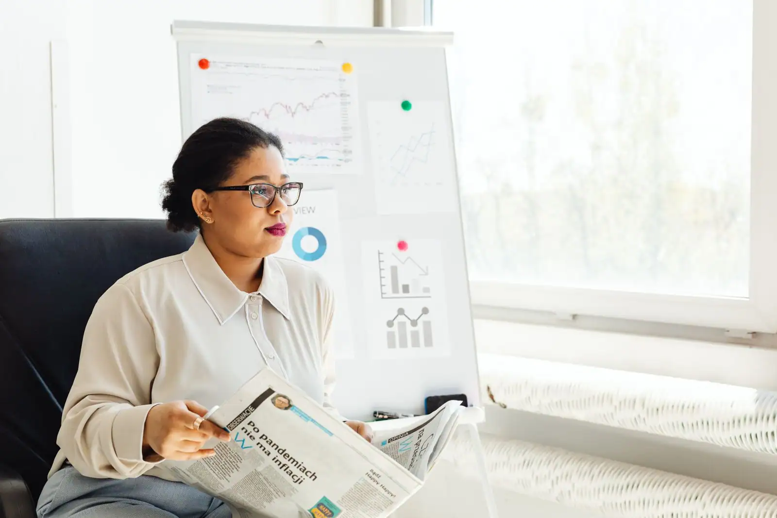 Femme lisant un journal économique devant un tableau de graphiques boursiers.
