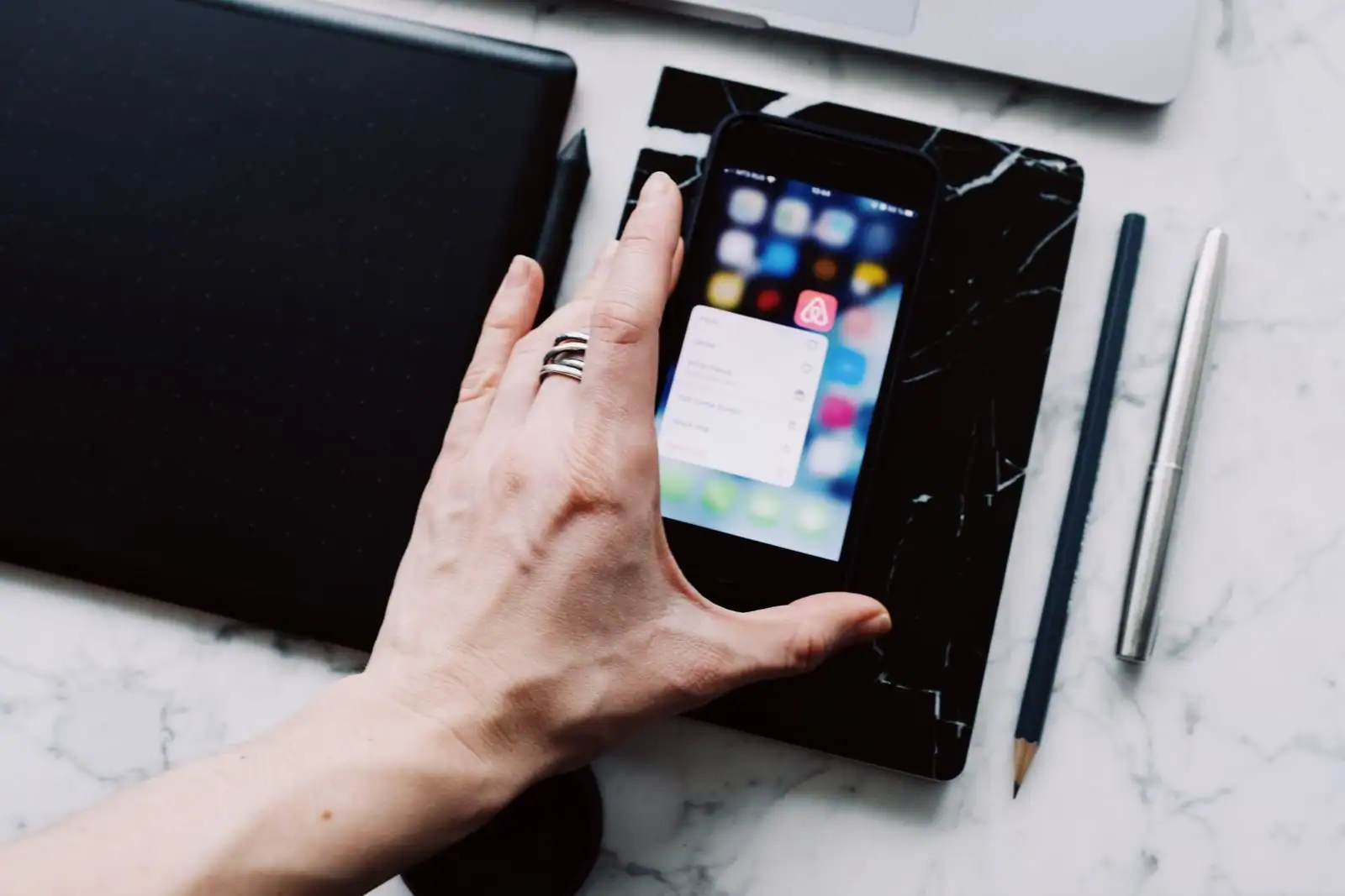 Une main saisit un smartphone affichant son écran d'accueil, posé sur un bureau avec une tablette graphique et des stylos.