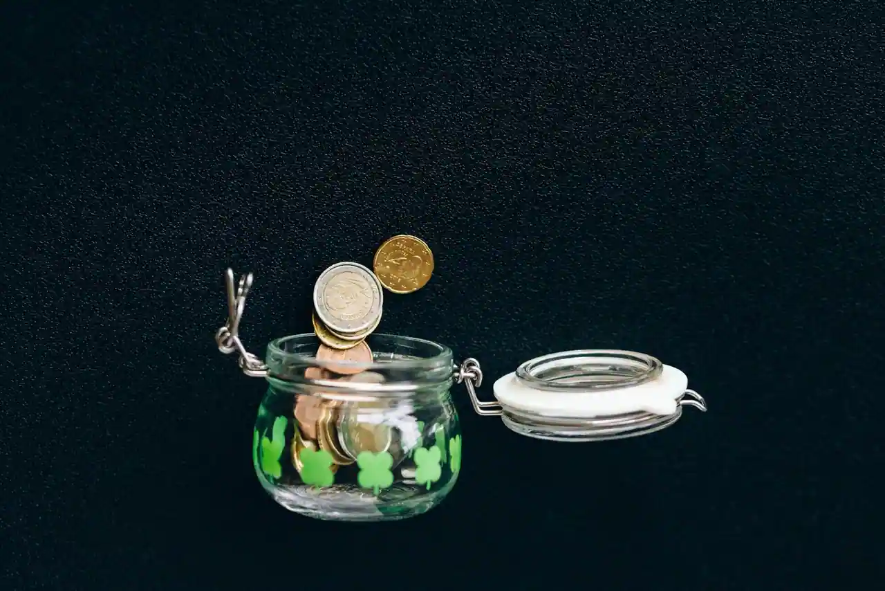 Bocal en verre rempli de pièces d'euros, avec quelques pièces posées sur le dessus, sur fond sombre.