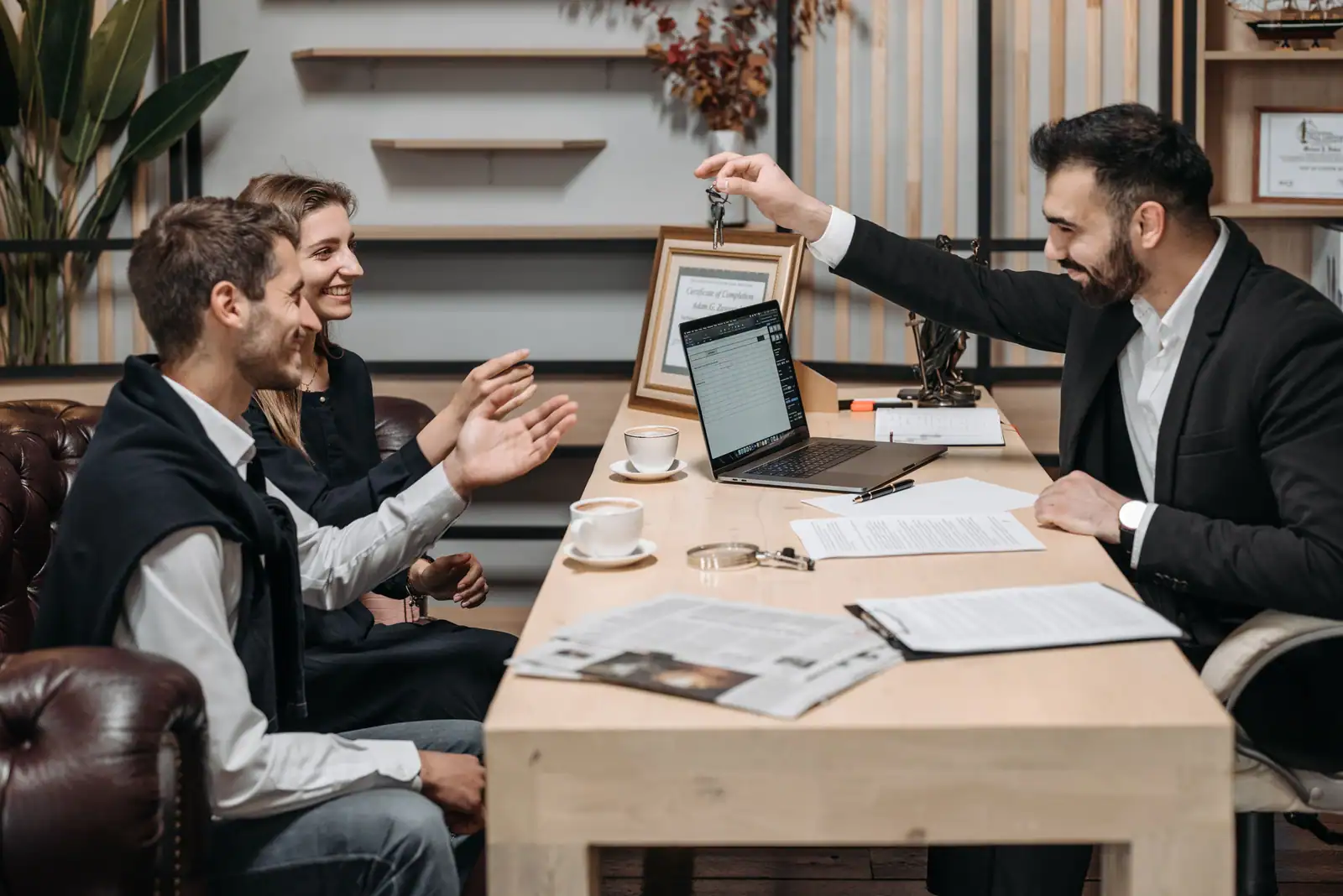 Un professionnel de l'immobilier tend les clés à un couple souriant lors d'un rendez-vous, contrat affiché sur l'ordinateur portable posé sur le bureau