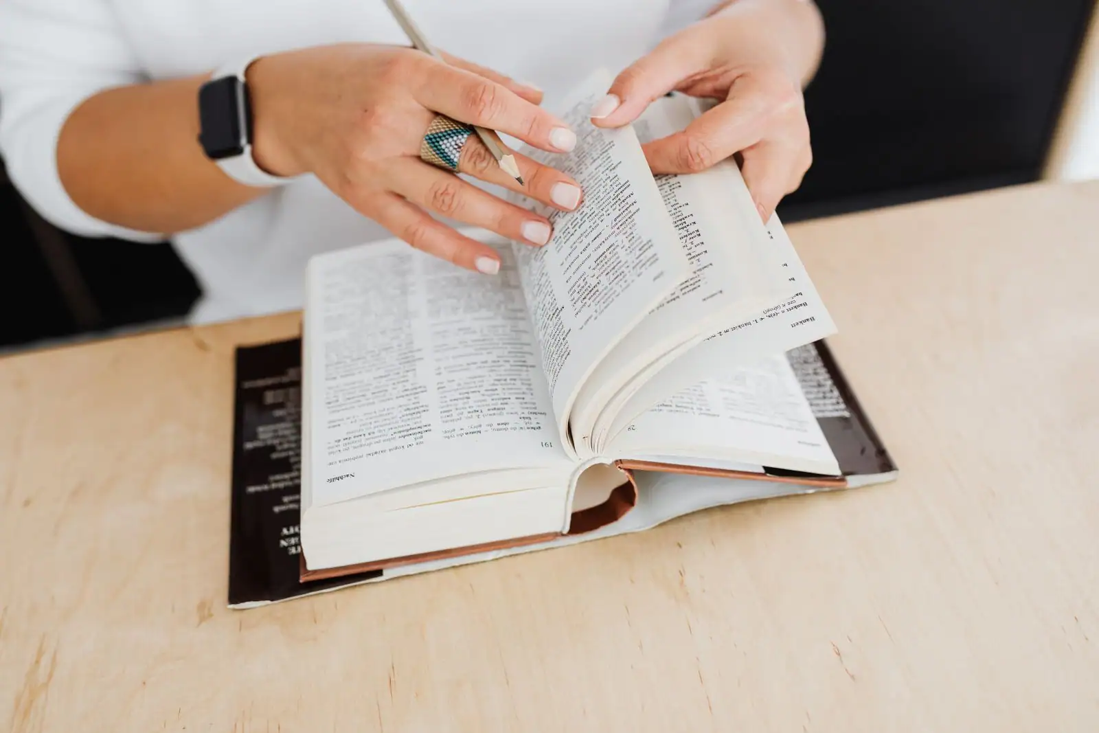 Mains feuilletant un dictionnaire ouvert, posé sur un bureau en bois clair.