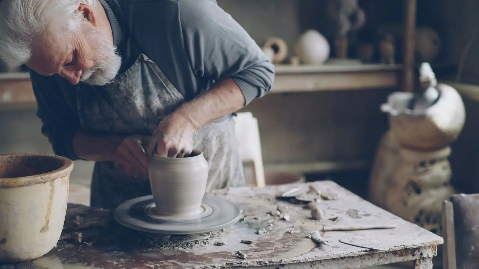 Homme âgé aux cheveux blancs façonnant un pot en terre au tour dans un atelier de poterie